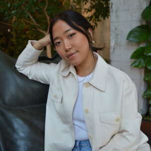 Headshot of Elisabeth Clarke smiling softly at the camera, wearing a cream button-up jacket, white top, and gold earrings, with one hand raised to their head, seated against a dark leather surface with greenery and a white brick wall in the background.