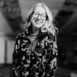 Black-and-white headshot of Elise Weber smiling, wearing glasses and a patterned fleece zip-up jacket, with a blurred outdoor background.
