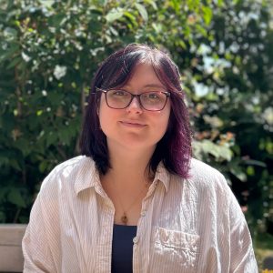 Headshot of Kathleen Mascia smiling outdoors, wearing glasses, a white striped button-down shirt, and a gold necklace, with green foliage in the background.