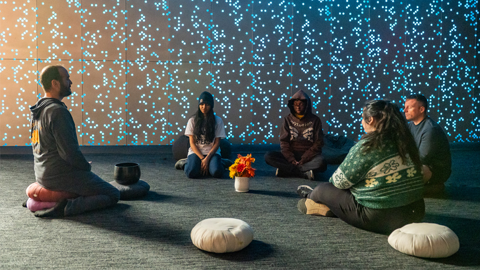 Hoffner sits on a pillow on the floor facing four seated students. In the background, the center's wall twinkles with dozens of tiny lights. The lighting in the center is muted and peaceful.