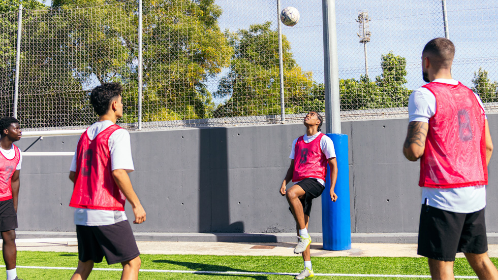 Four college students stand across from each other practicing soccer skills — one is bouncing the ball off his head.