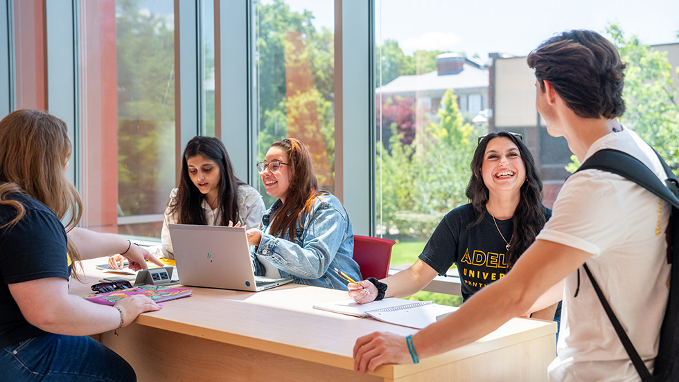 A group of male and female students gather around a bench, studying. chatting and collaborating.