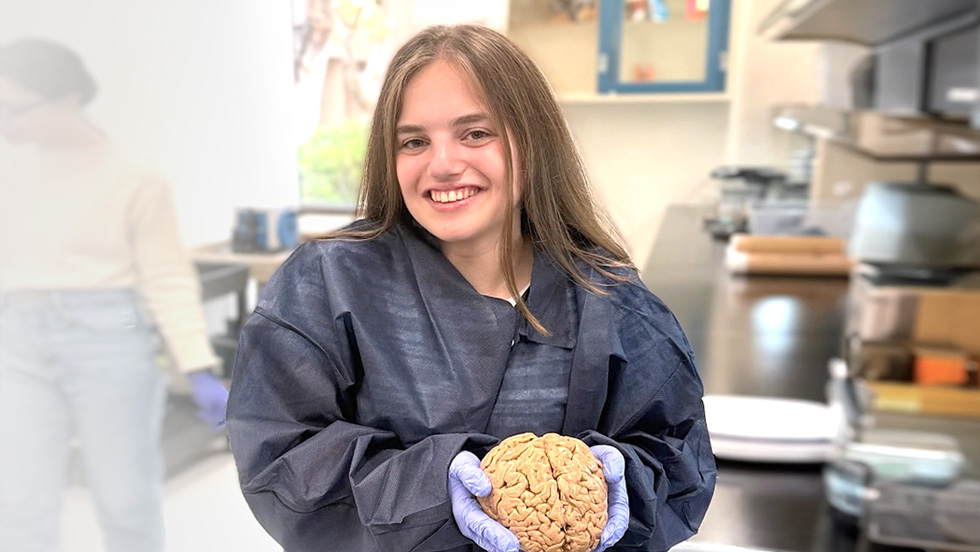 A young woman with long brown hair smiles while wearing a dark blue lab gown and purple gloves. She is holding a human brain specimen in a laboratory setting with lab equipment visible in the background.