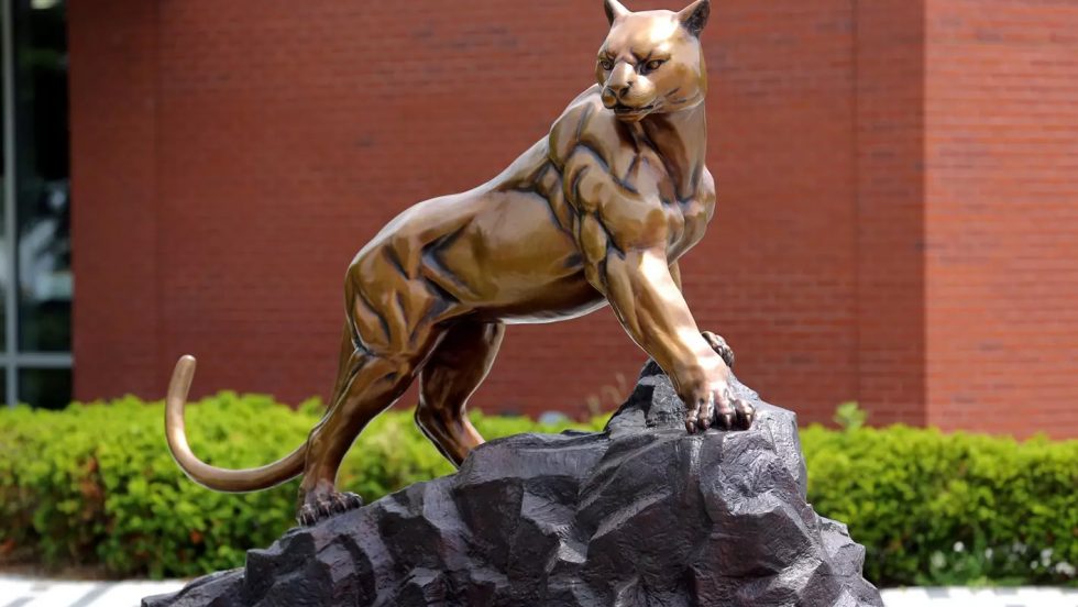 A bronze statue of a muscular panther perched on a jagged rock, set against a brick building and green shrubbery.