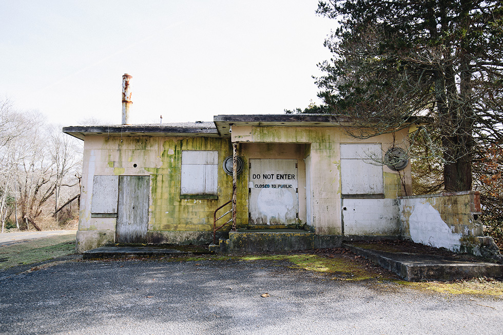 A small, abandoned one-story building with moss-covered walls and boarded-up windows, featuring a "Do Not Enter" sign on the main door.