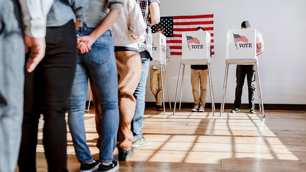 A line of people waiting to vote at a polling station. In the background, three individuals stand in voting booths labeled "VOTE" beneath a large American flag hanging on a white wall.