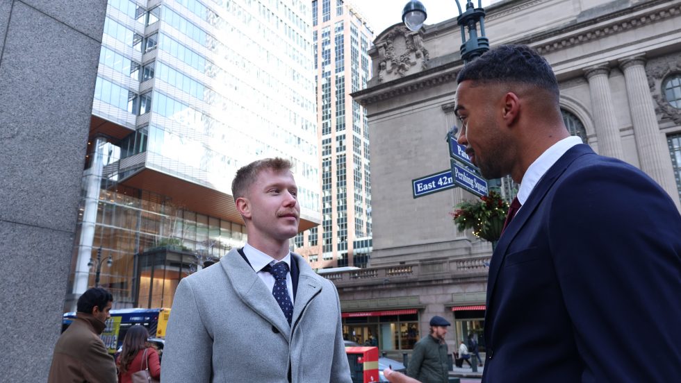 Two men in professional attire stand on a busy street corner in New York City, engaged in conversation. 