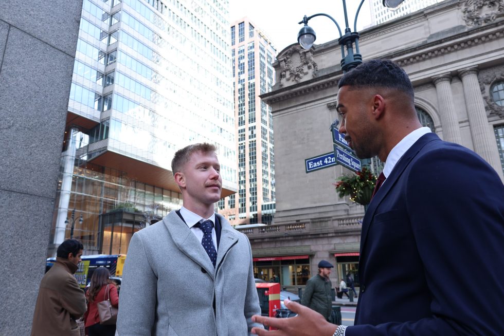 Two men in professional attire stand on a busy street corner in New York City, engaged in conversation. 