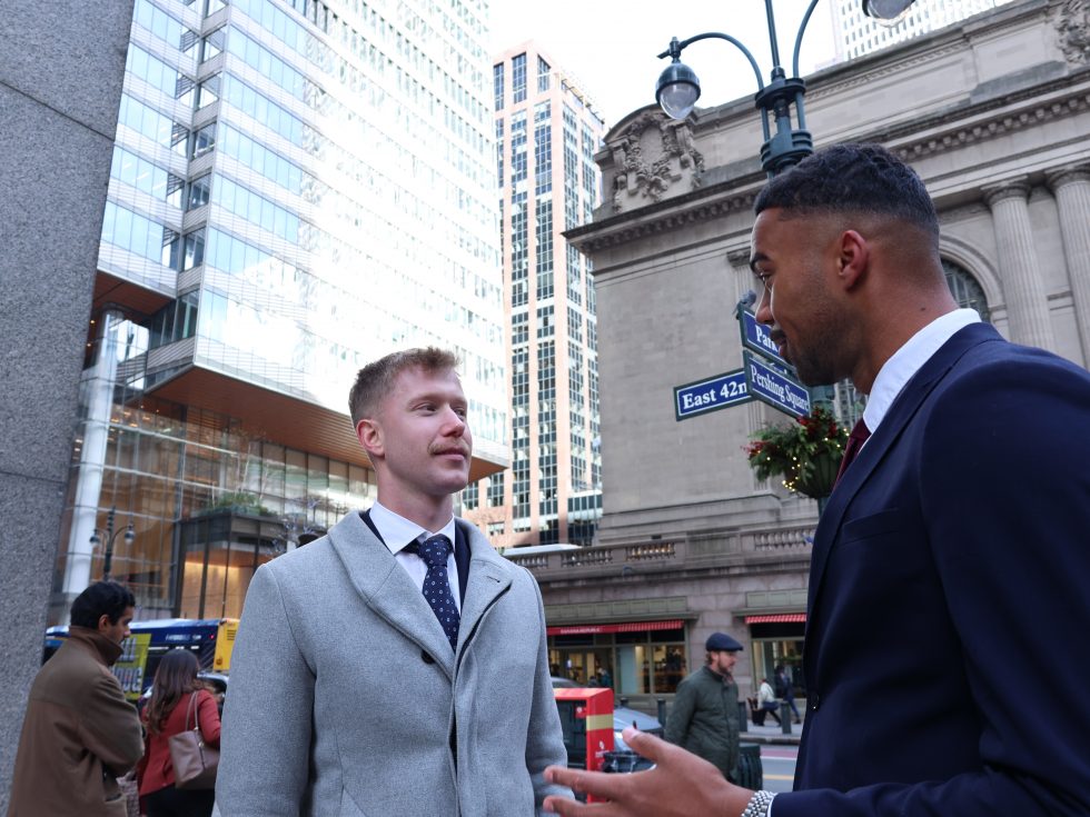 Two men in professional attire stand on a busy street corner in New York City, engaged in conversation.