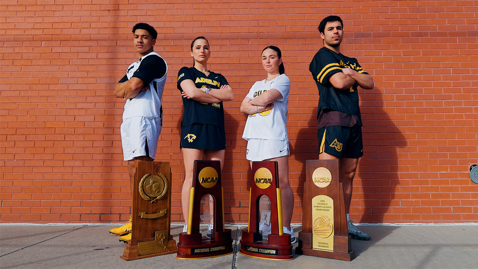The student-athletes, wearing their teams' uniforms, stand with their arms folded before a brick wall and behind a quartet of national championship trophies.