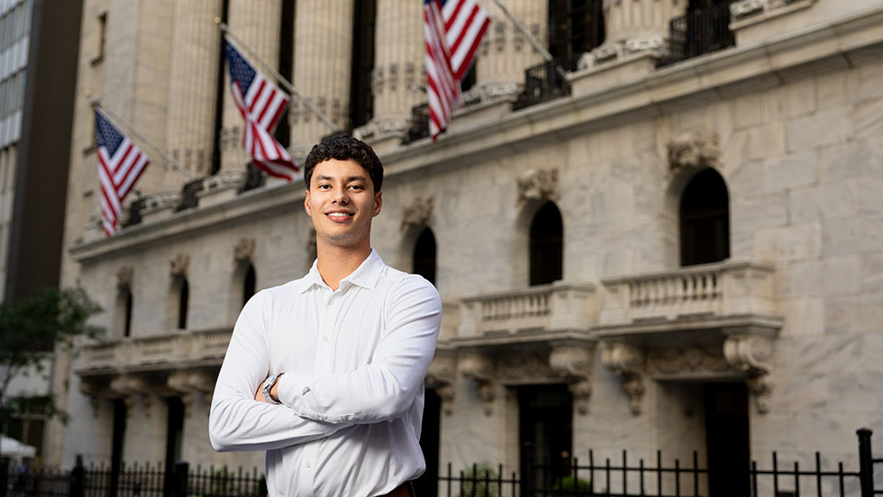 A male student in a collared shirt standing in a city setting with his arms crossed.