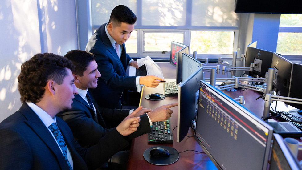 Three male business students in professional suits work collaboratively in a high-tech finance lab.