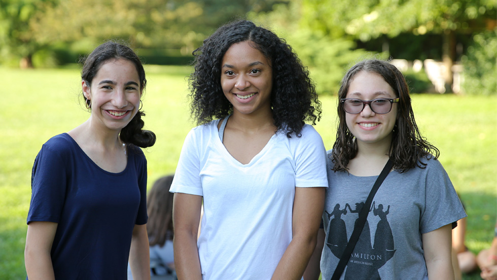 Three young women smiling outdoors in a park setting. From left to right: a young woman in a navy blue t-shirt with dark hair pulled back; a young woman in a white v-neck t-shirt with curly dark hair; and a young woman in a gray Hamilton t-shirt wearing glasses, with a crossbody bag. Green trees and grass are visible in the background.