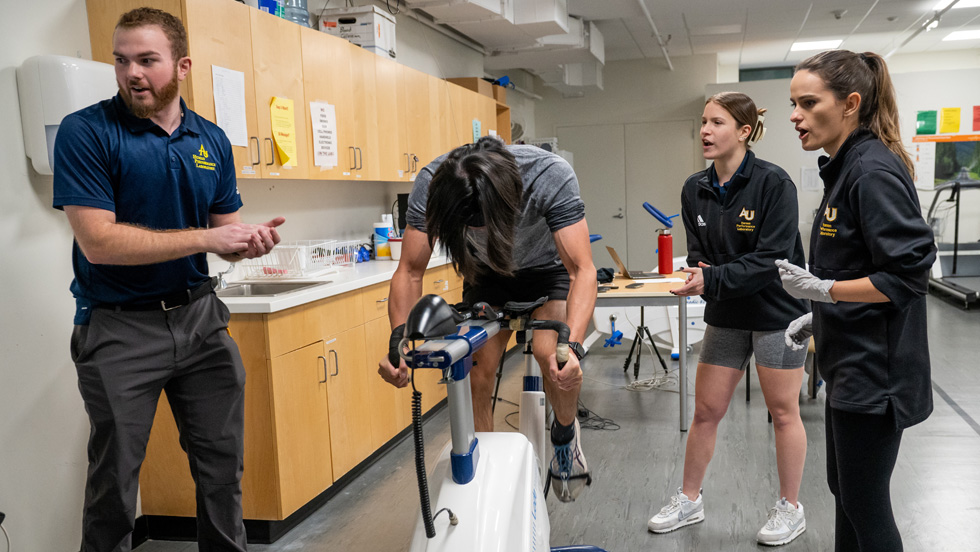 A professor and two graduate exercise science students observe a client on a stationary bicycle, and they seem to be encouraging him to keep pedaling.