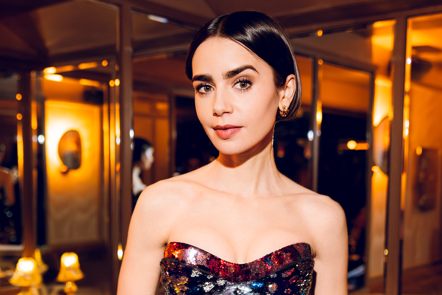 A woman with short dark hair poses indoors wearing a strapless, multicolored sequined dress.