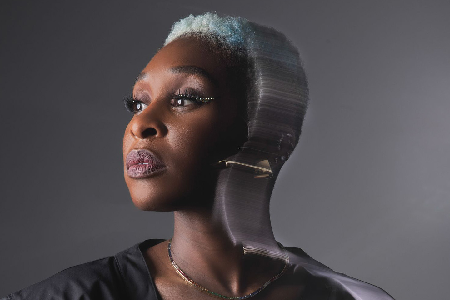 A studio portrait of a woman with short, light-colored hair wearing a black top. She gazes to the side against a gray background.