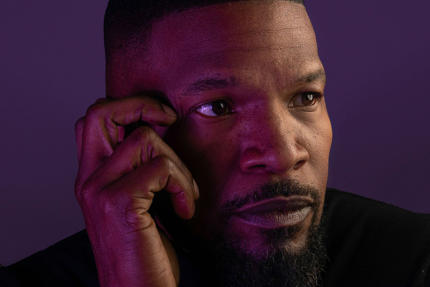 A close-up portrait of a man with a short haircut and trimmed beard resting his hand against his face.