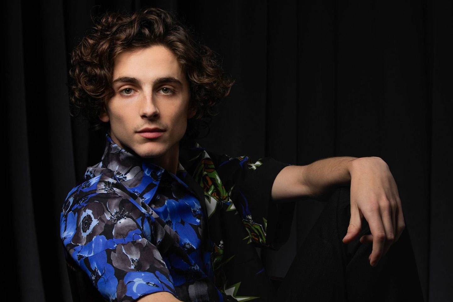 A studio portrait of a man with curly hair wearing a blue floral-patterned shirt