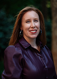 A women with long brown hair and a shiny satin blouse smiles warmly toward camera. She is Louise Geddes, PhD, is an expert on Shakespearean drama and performance who teaches English in the College of Arts and Sciences.