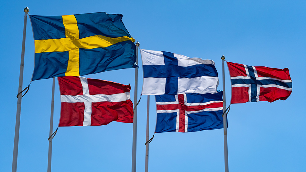 Five Nordic flags flying on poles against a clear blue sky: Sweden (blue with yellow cross), Denmark (red with white cross), Finland (blue with white cross), Iceland (blue with red and white cross), and Norway (red with blue and white cross).