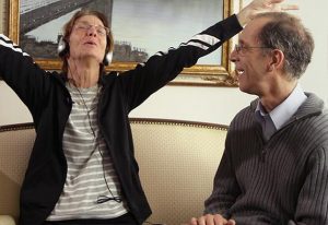 An older woman wearing headphones raises her arm expressively while listening to music, seated on a sofa next to a smiling man who watches her with delight. A framed painting of a bridge hangs on the wall behind them.