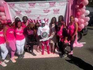 A group of women pose together in front of a step-and-repeat banner for the Spring Forward Sweat Fete Pink Awareness Health & Fitness Event, surrounded by pink and white balloons. Most participants wear bright pink athletic wear; one woman in the center holds a Spring Forward Consulting, LLC award or plaque.