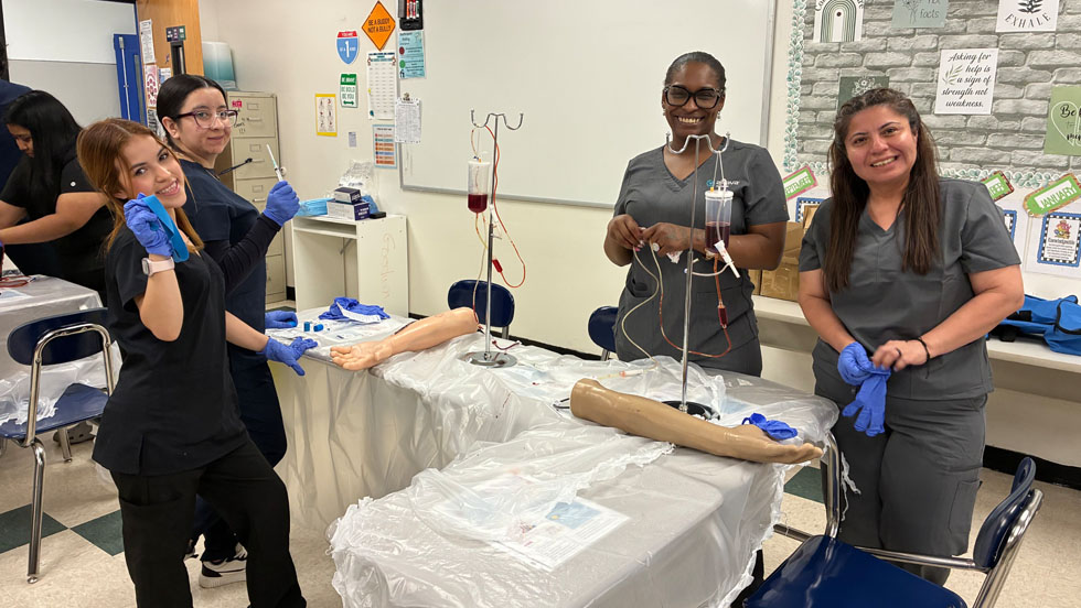 Four students in gray and black scrubs smile at the camera while practicing phlebotomy skills on medical training arm models with IV equipment in a classroom.