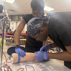 Two students wearing blue latex gloves practice a phlebotomy technique on a medical training arm model connected to IV tubing in a classroom setting.