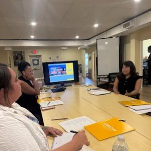 A group of people seated around a conference table during a meeting, with Adelphi University branded yellow folders on the table and a monitor displaying a virtual meeting in the background.