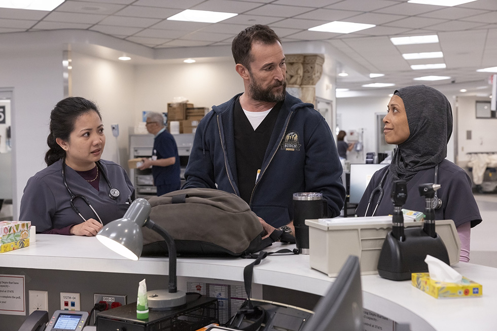 Three healthcare workers—two nurses and a male colleague—stand at a clinical station desk, engaged in discussion about patient care or administrative matters.