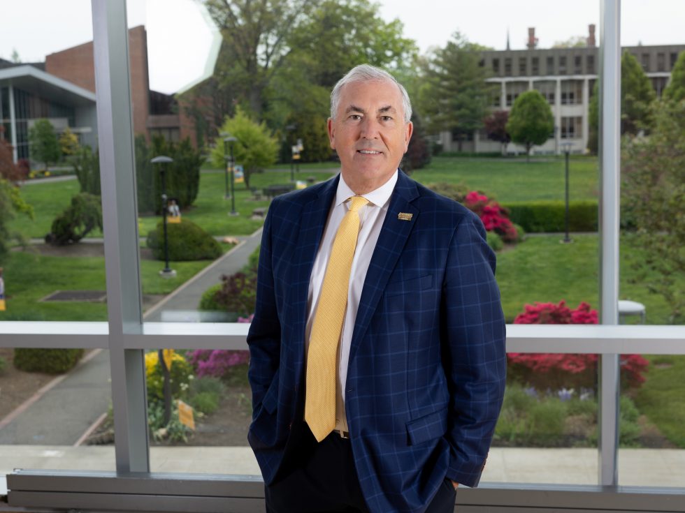 A man standing in business suit in front of a window with a landscape background of trees, a path and bushes.
