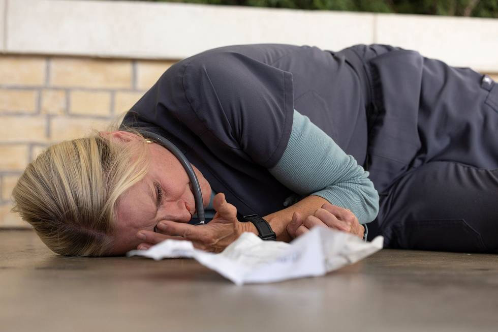 A healthcare professional wearing a stethoscope and dark jacket lies on the ground in a recovery position during a first aid or emergency response training exercise.