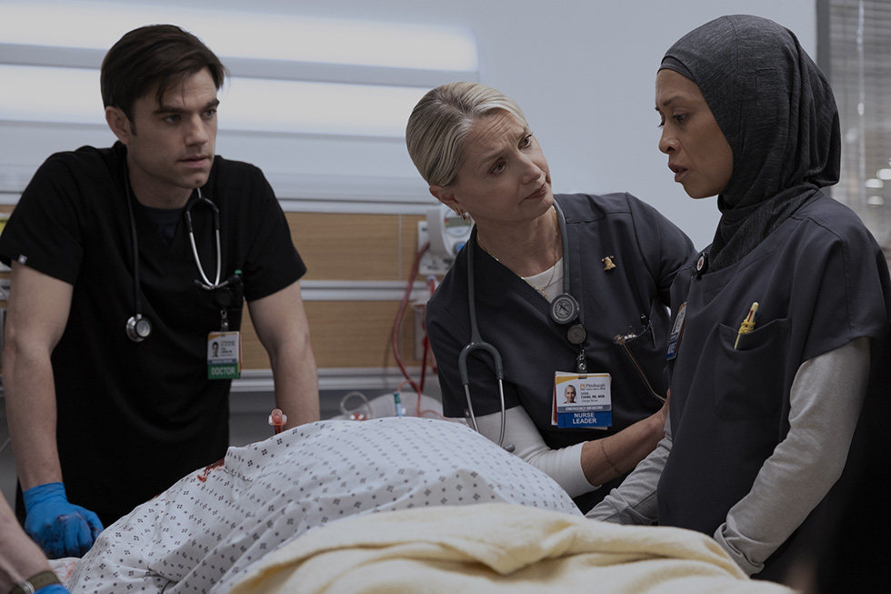 A senior nurse instructor in dark scrubs speaks with two nursing students at a bedside, pointing to and discussing a patient simulation mannequin's condition.