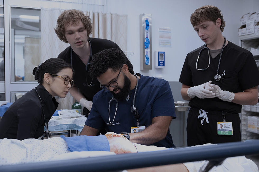 Four nursing students in scrubs and stethoscopes gather around a patient simulation mannequin in a clinical lab, examining and discussing treatment procedures.