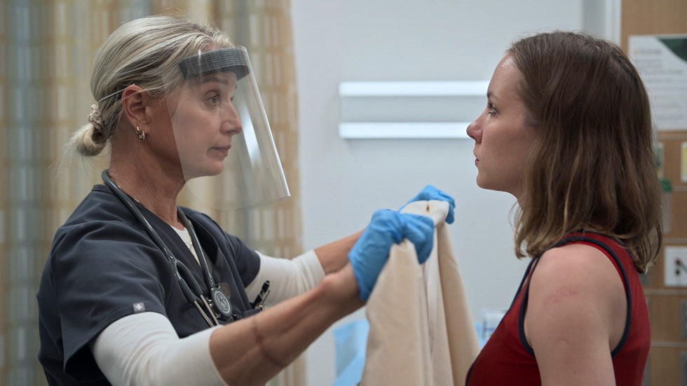 A healthcare worker wearing a face shield and blue gloves administers a vaccine or medical injection to a patient's arm in a clinical setting.