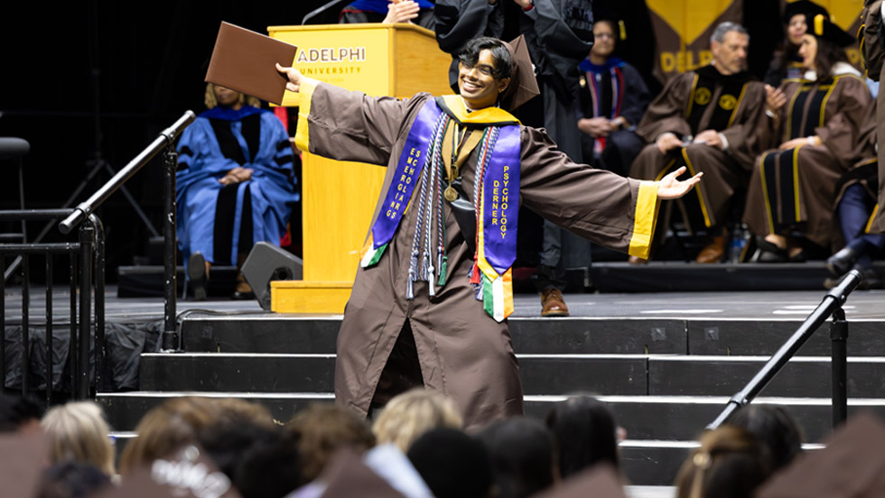 Student wearing Adelphi graduation attire with his arms out and smiling on stage at the Commencement ceremonies. 