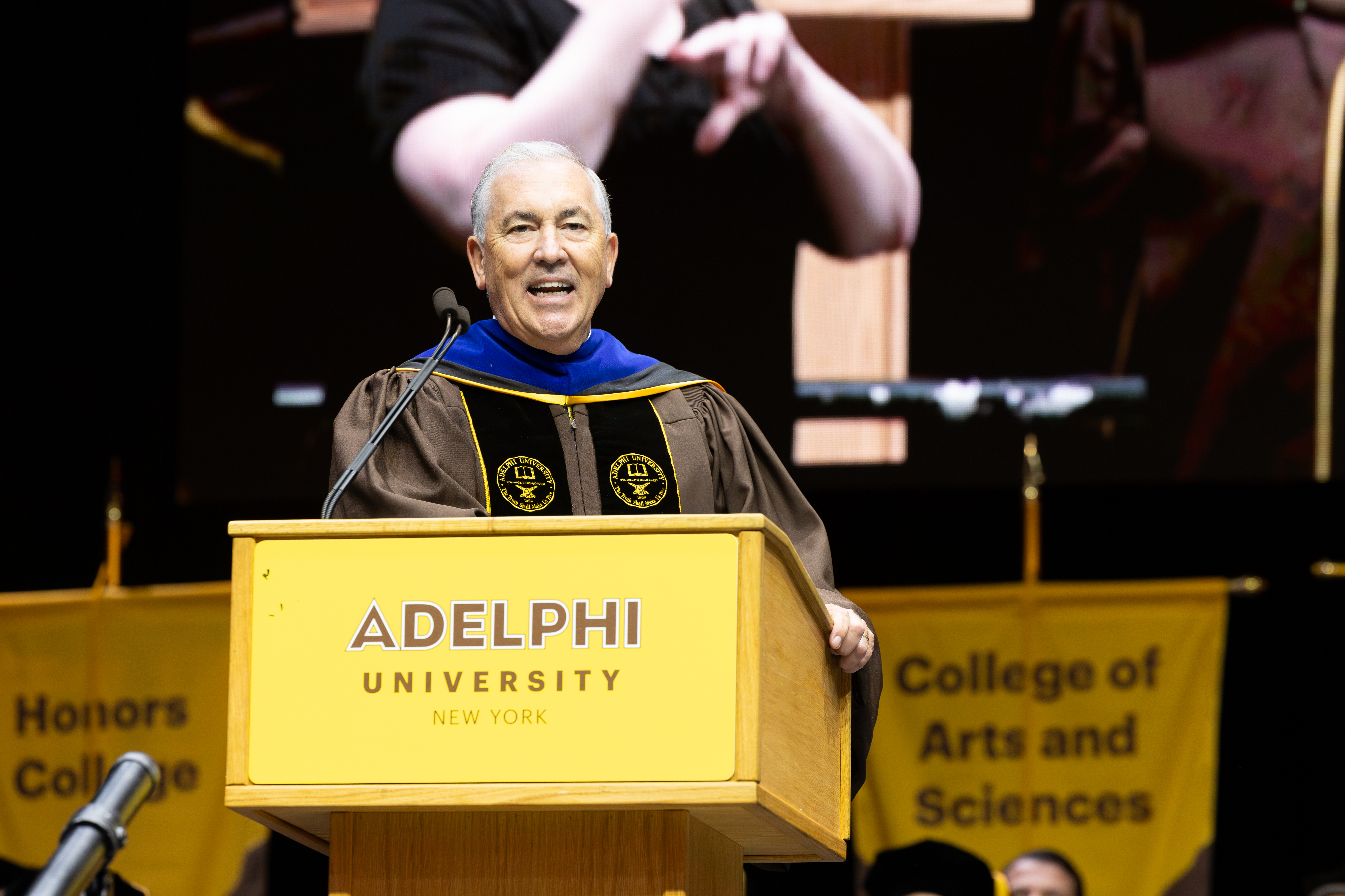 A man wearing graduation regalia speaking at a podium on a stage.