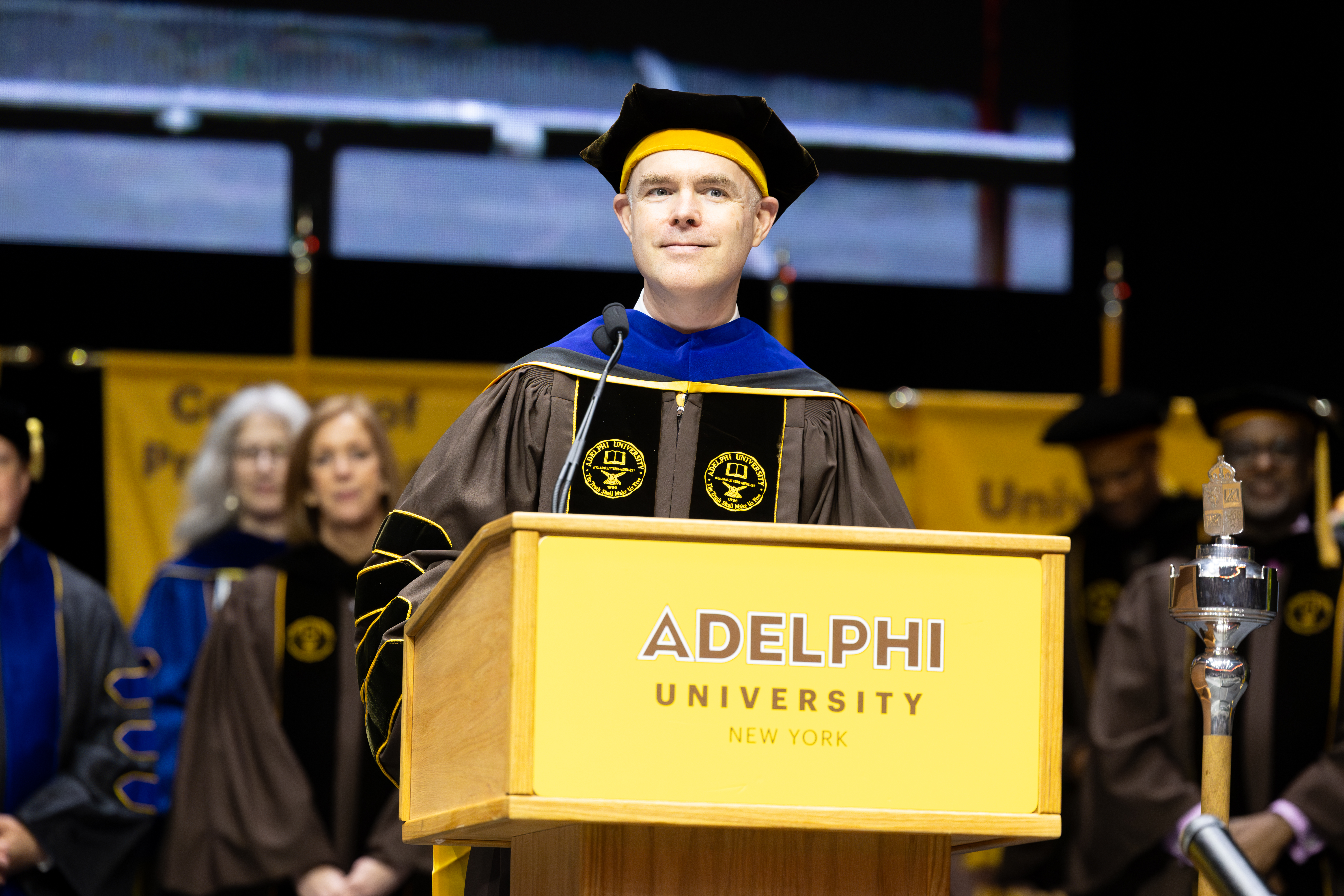 A man wearing graduation regalia speaking at a podium on a stage.