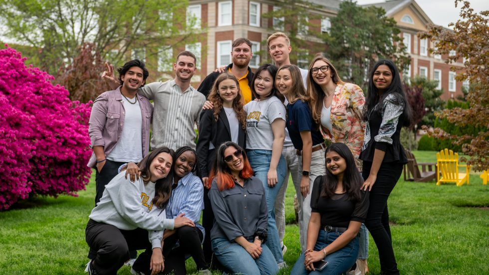 A group of a dozen students pose on Adelphi's campus in springtime next to a large azalea in full flower.