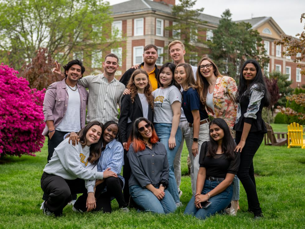 A group of a dozen students pose on Adelphi's campus in springtime next to a large azalea in full flower.