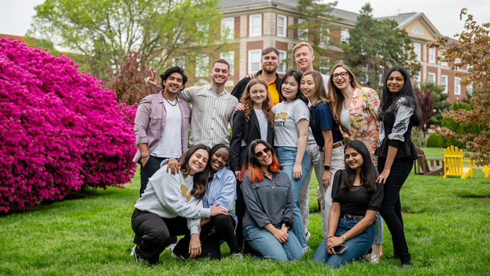 A group of a dozen students pose on Adelphi's campus in springtime next to a large azalea in full flower.