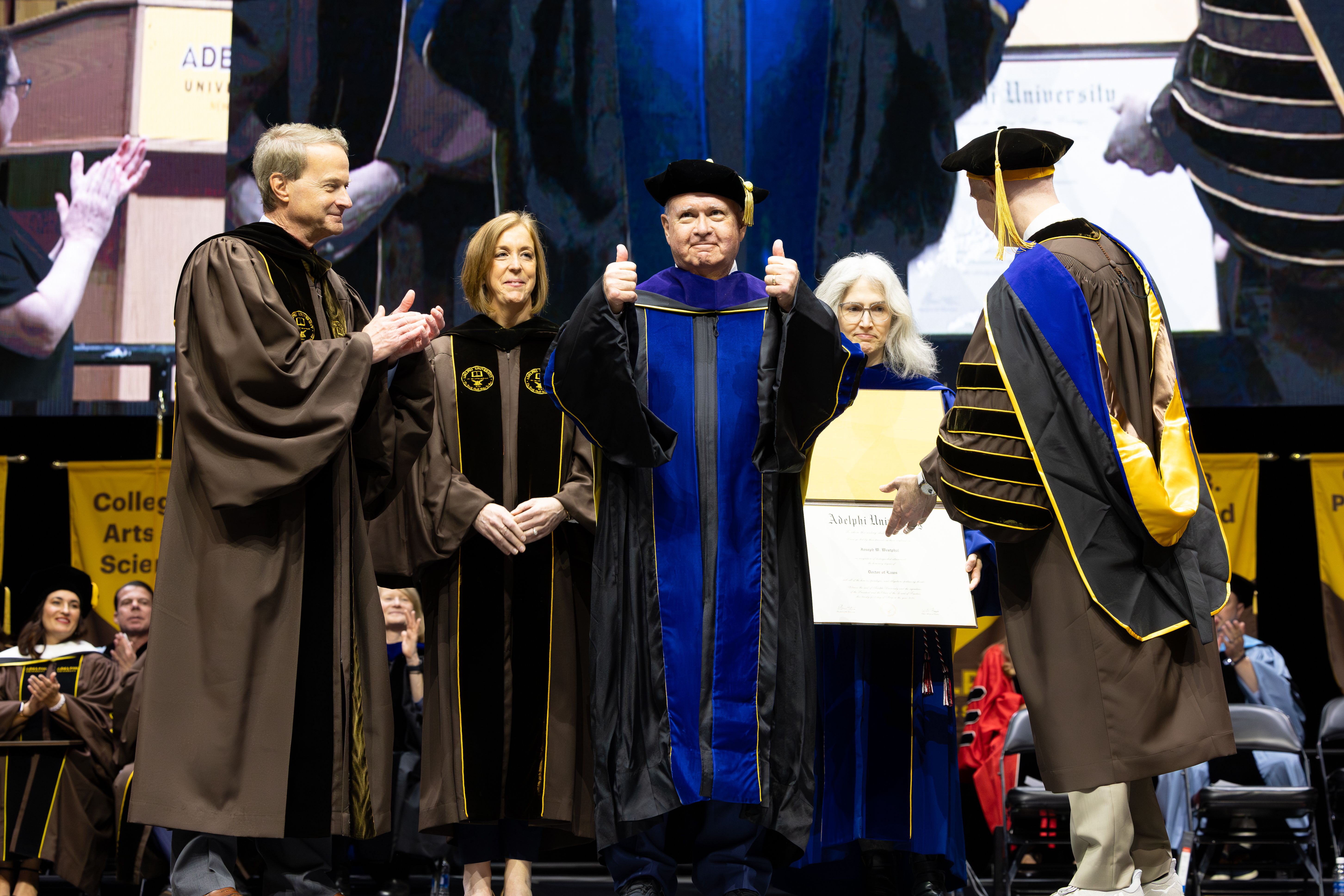 A man in graduation regalia on stage surrounded by other graduation officials being given an honorary degree.