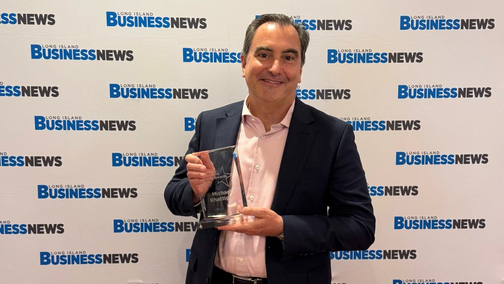 A man in business attire holding a glass award in front of a step-and-repeat that says “Long Island Business News.” 