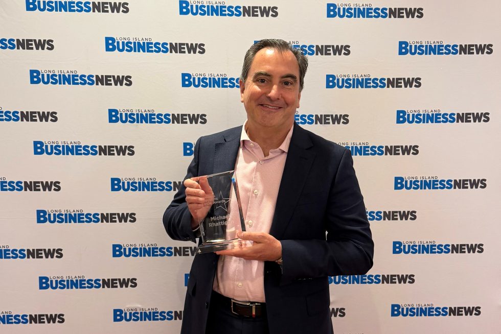 A man in business attire holding a glass award in front of a step-and-repeat that says “Long Island Business News.” 