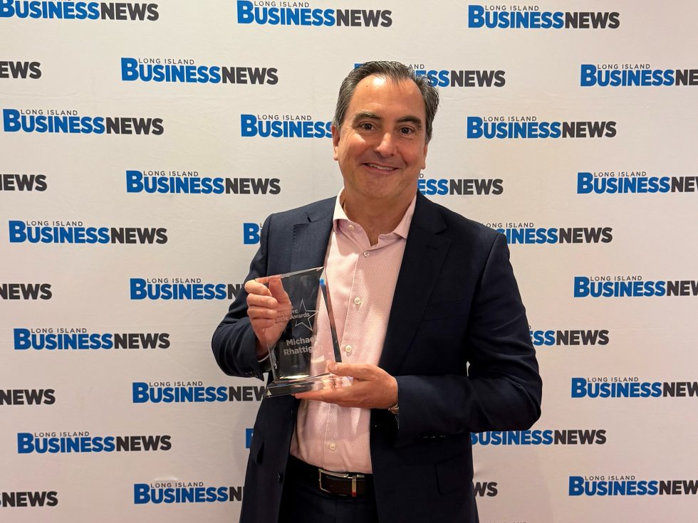 A man in business attire holding a glass award in front of a step-and-repeat that says “Long Island Business News.” 
