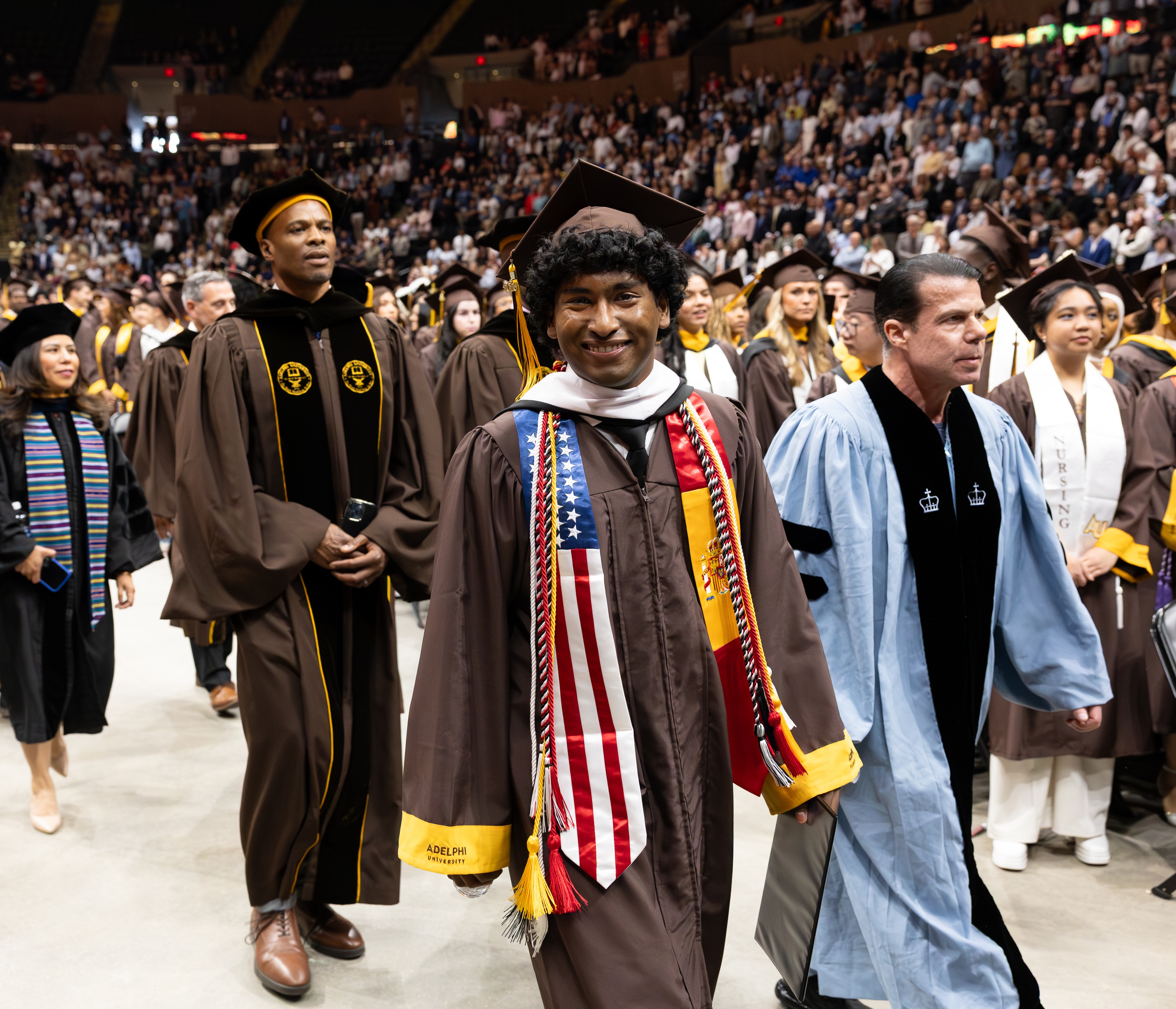 A male student wearing graduation attire walking happily to the commencement stage amongst faculty members.