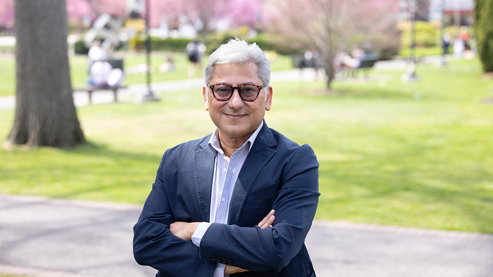 A man wearing a suit and glasses, Adelphi's first Fulbright-Scholar-in Residence, poses in front of the beautiful Adelphi campus in spring time.