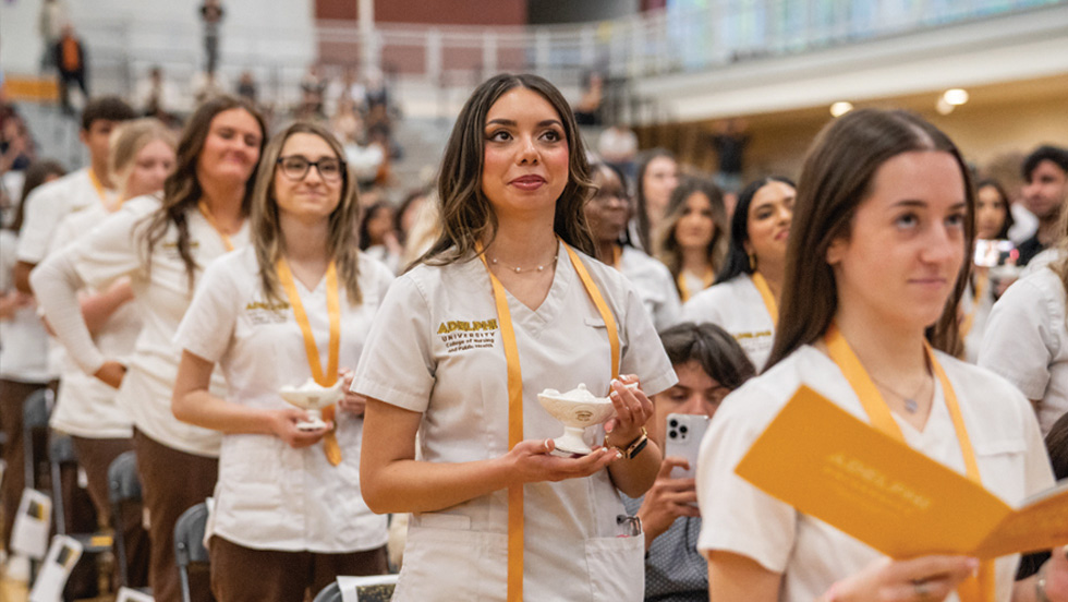 Group of nursing graduates wearing white uniforms and gold honor cords stand together in a gymnasium during a pinning ceremony, holding candles and commemorative items as they celebrate their transition to professional practice. 