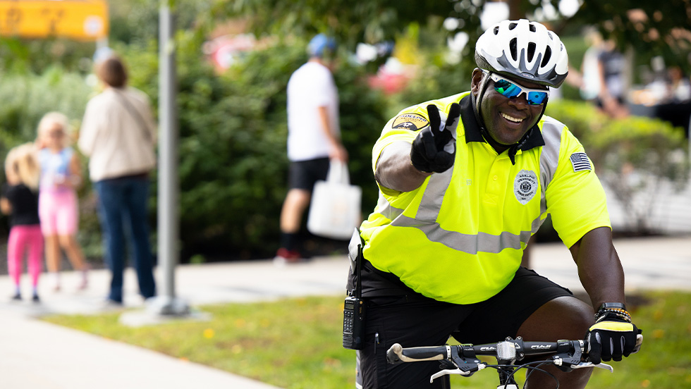 A police officer rides by on a bicycle through the campus, giving a friendly wave and a peace sign. 