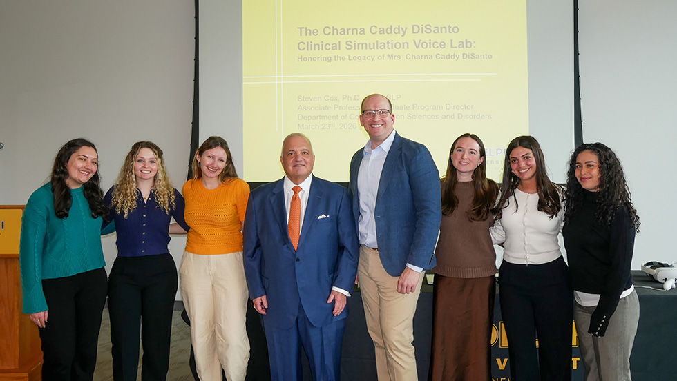 A group of graduate students, along with donor Tony DiSanto MBA '88 and Steven Cox, PhD, associate professor, communication sciences and disorders, gather in front of a screen announcing the new Charna Caddy DiSanto Clinical Simulation Voice Lab.
